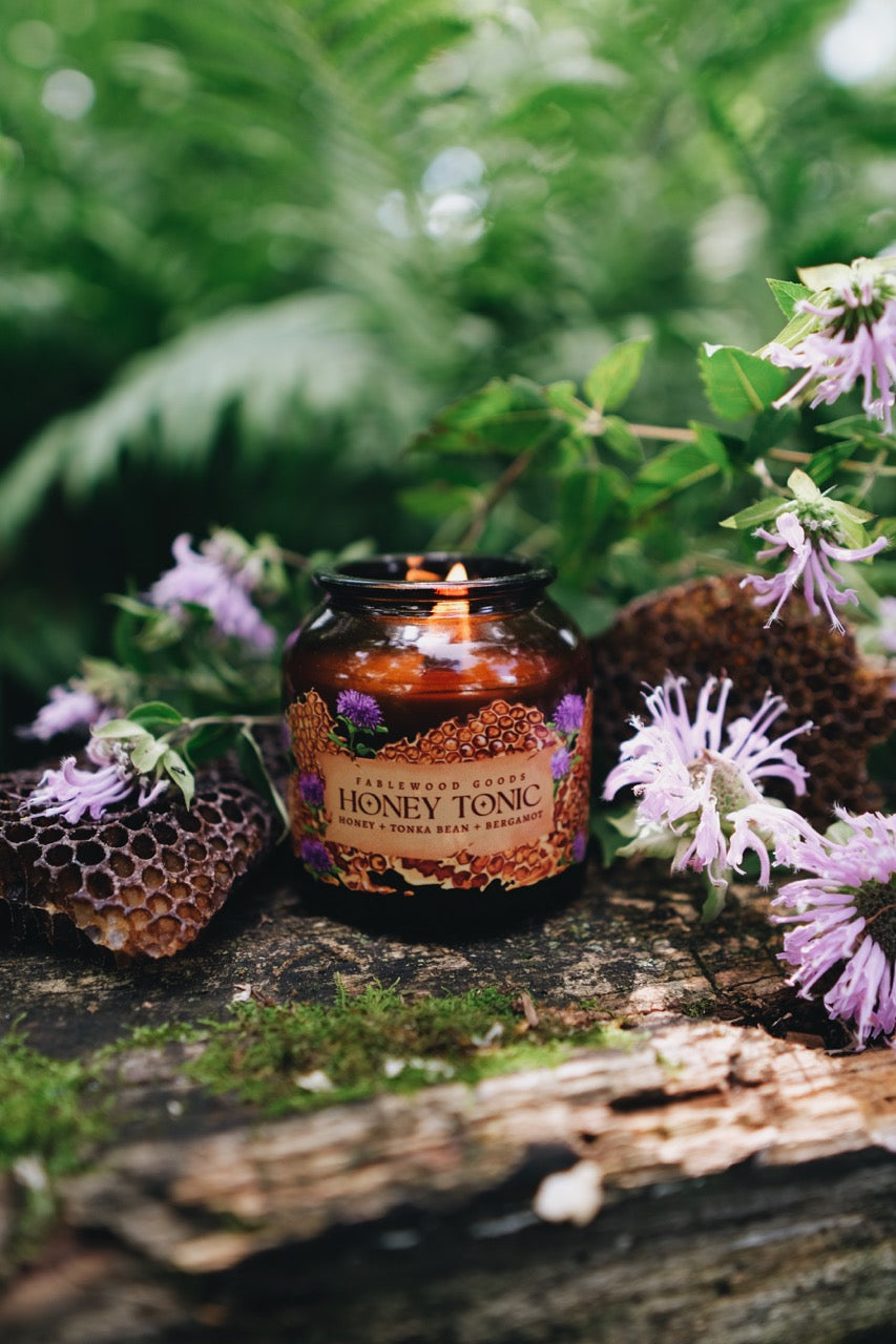 Candle labeled 'Honey Tonic' surrounded by flowers and a honeycomb on a natural background