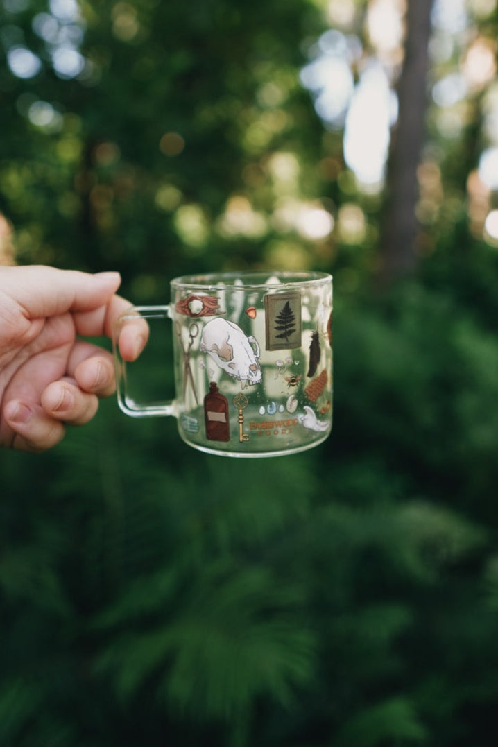 Hand holding a glass mug with illustrations against a blurred green outdoor background